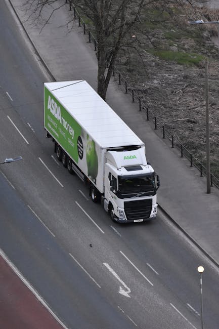 An aerial view of a white ASDA delivery truck parked on a paved urban street, adjacent to a sidewalk lined with a metal railing and a row of leafless trees. The truck features a large green ASDA Money