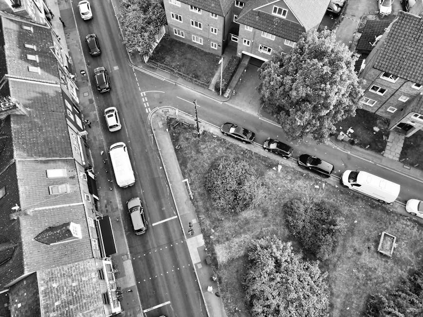 A narrow urban street view showing a wet pavement surface with a white van parked further along the road. On the left side, there are multi-storey buildings with brick and glass facades, some with bal